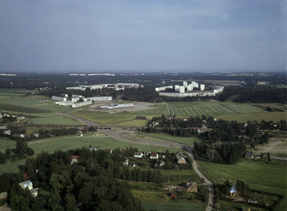 Ilmakuva: etualalla on peltoja ja pientaloja, kauempana vaaleita kerrostaloja metsäisten alueiden lomassa.